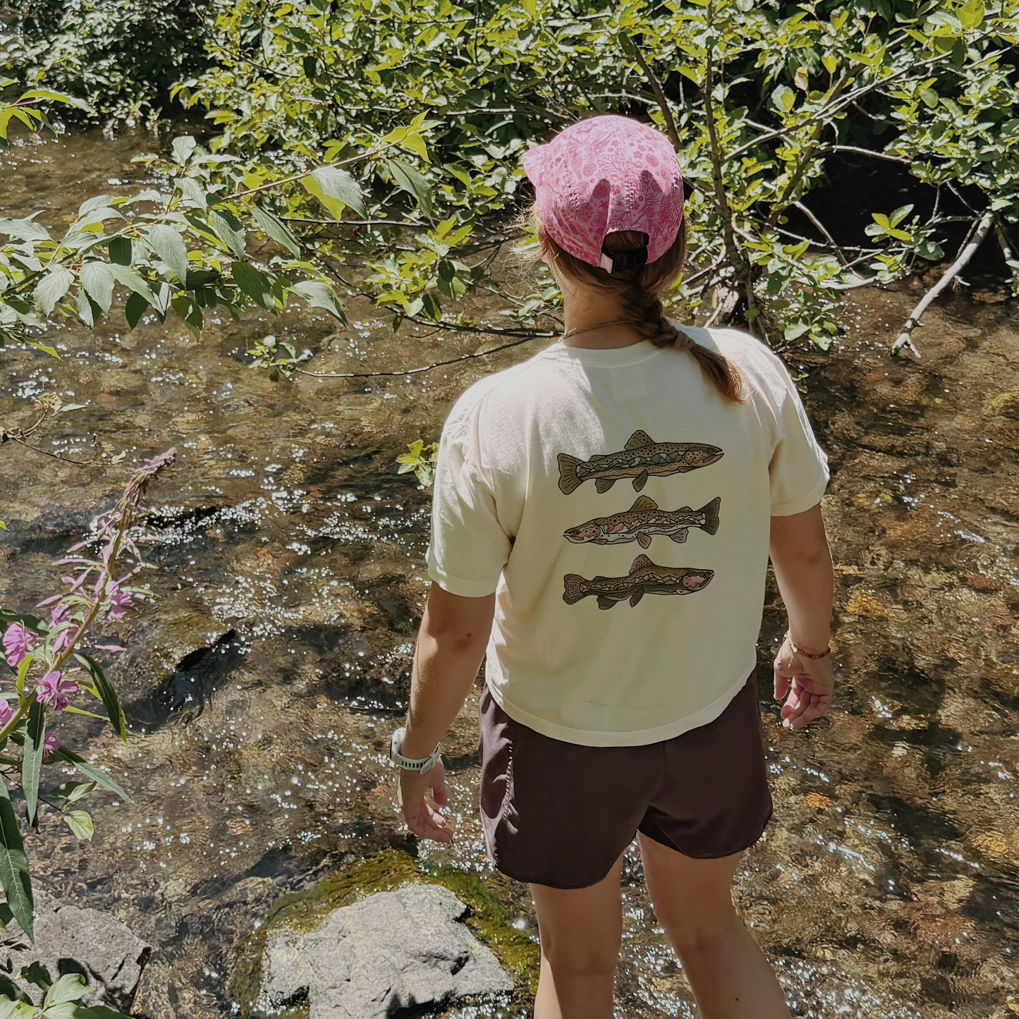 A person standing in a shallow stream, viewed from behind, wearing a cream t-shirt with three fish graphics on the back, brown shorts, and a pink cap, surrounded by green foliage and water.