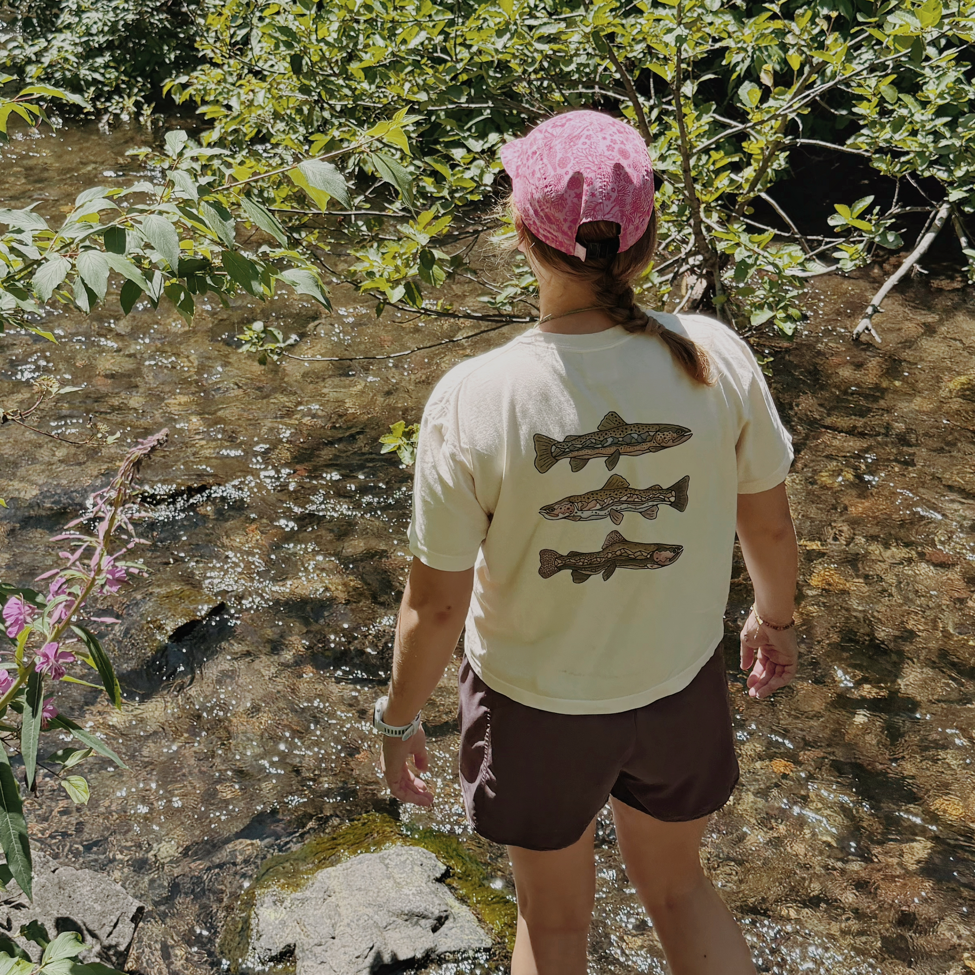 A person standing in a shallow stream, viewed from behind, wearing a cream t-shirt with three fish graphics on the back, brown shorts, and a pink cap, surrounded by green foliage and water.